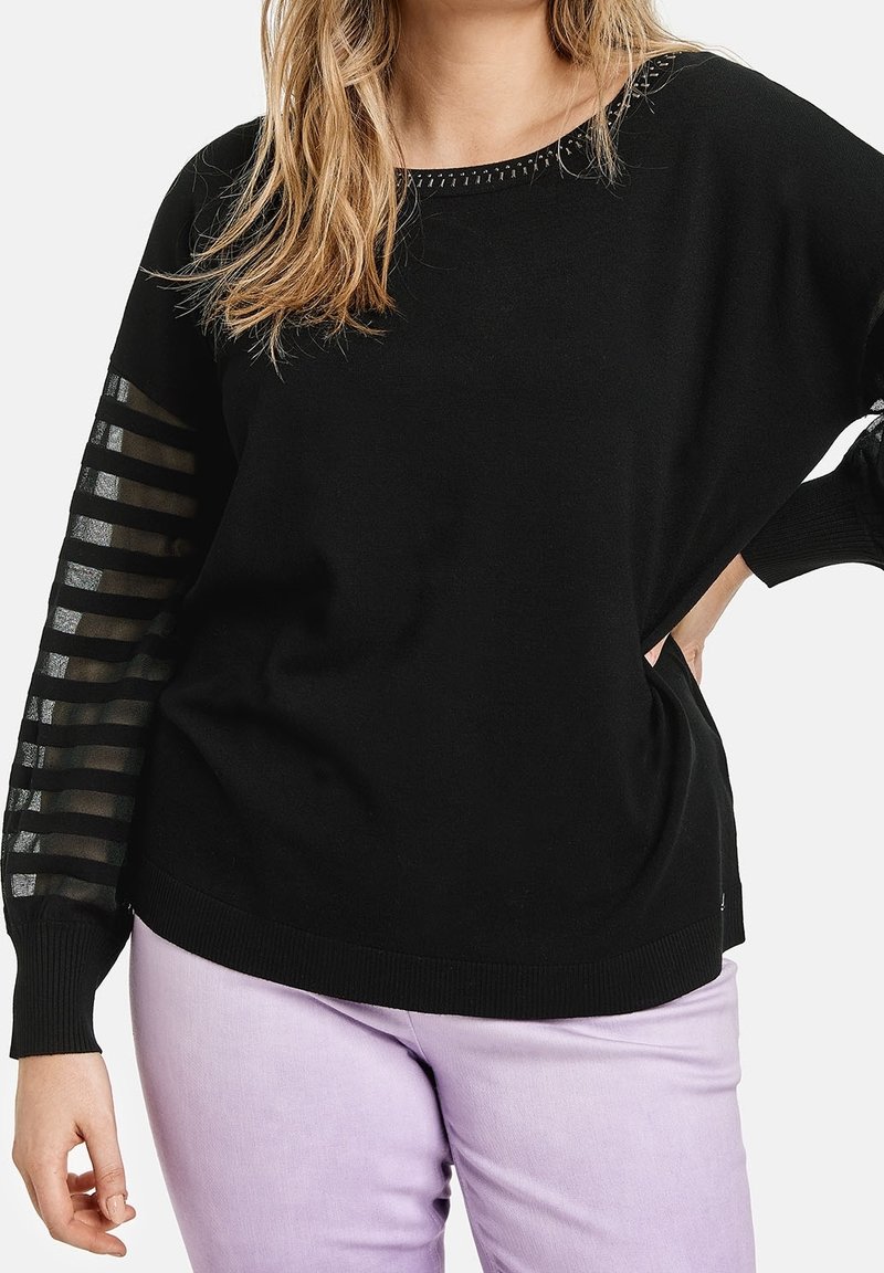 Femme portant un pull noir avec des manches rayées transparentes et un pantalon lavande, posant avec une main sur la taille devant un fond blanc.