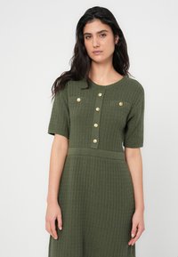 Πλεκτό ελιάς πράσινο φόρεμα με κοντά μανίκια, υφασμάτινη υφή, στρογγυλή λαιμόκοψη και χρυσά κουμπιά με αντίστοιχες τσέπες στο στήθος.