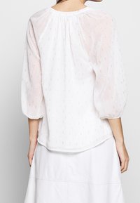 Blusa bianca in tessuto morbido e trasparente, con scollatura arricciata e maniche a sbuffo 3/4. Delicati accenti metallici creano un motivo testurizzato.