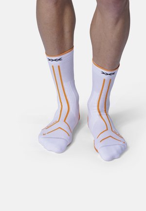 Twee benen met witte sportsokken met oranje strepen en zwarte logo's op een effen lichte achtergrond, staand met voeten dicht bij elkaar.