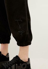 Zwarte velours joggers met elastische boorden, voorzien van een getextureerd geborduurd ontwerp op het linkerbeen. Gepaard met zwarte sneakers.
