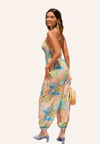 Une combinaison à motif paisley multicolore avec un buste froncé, des jambes larges et des bretelles réglables, assortie de sandales à talons blocs bleus.