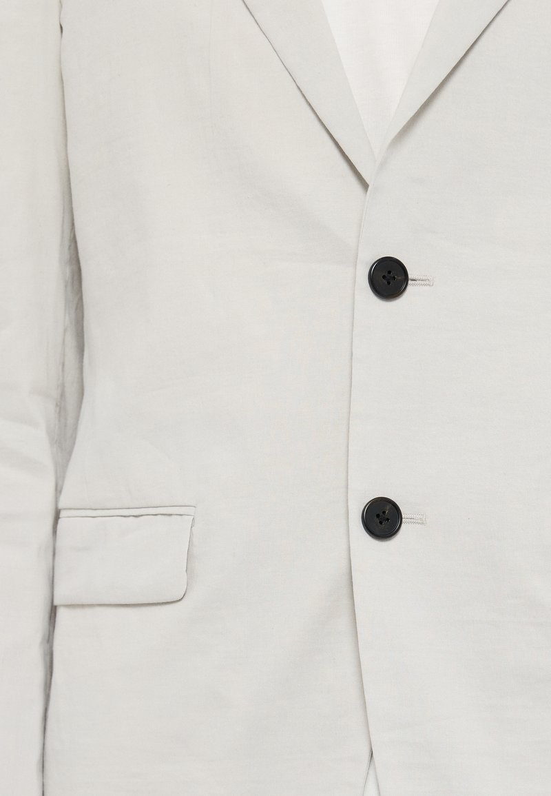 Blazer di lino grigio chiaro con vestibilità slim, dotato di due bottoni neri e una trama sottile. Include una tasca sul petto e rever a punta.