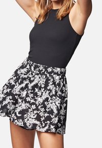 Žena v bezramenném černém žebrovaném topu a černé sukni s květinovým vzorem a elastickým pasem, pózující s rukama zdviženýma.