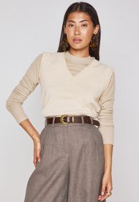 Maglione beige a coste con scollatura a V indossato sopra una maglietta a maniche lunghe beige chiaro, abbinato a pantaloni marrone testurizzati a vita alta e una cintura marrone.