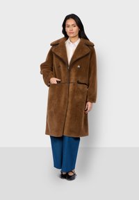 Cappotto in finta pelliccia marrone con ampi risvoltini, bottoni doppi e tasche laterali. Abbinato a pantaloni a gamba larga blu e scarpe nere.