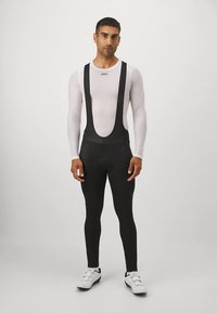 Črne kolesarske hlače z naramnicami iz raztegljive tkanine, z nastavljivimi trakovi in podloženim sedežem. Nošene čez oprijet bel dolg rokav.