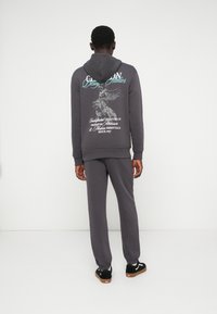 Hoodie cinza com uma impressão gráfica nas costas, combinado com calças a condizer e ténis pretos. Tecido macio com punhos e cintura canelados.