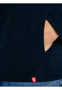 Prawa dłoń w kieszeni ciemnogranatowej kurtki polarowej, powyżej paska niebieskich dżinsów widoczna czerwona metka "BIG STAR".