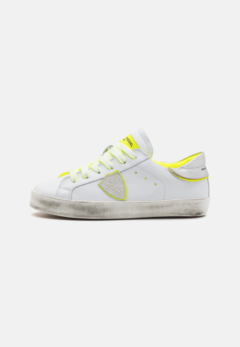 White leather sneaker with neon yellow accents, round toe, flat laces, textured sole, logo patch on side, and contrasting silver heel detail.