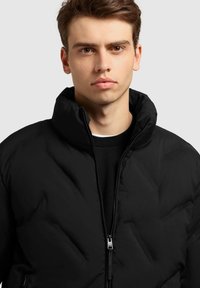 Veste matelassée noire avec col montant, fermeture éclair et détails de couture visibles. Portée sur une chemise sombre, mettant en valeur une texture lisse.