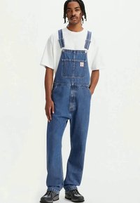 Mlad moški stoji oblečen v modre denim overale, belo preveliko majico in črne superge na enobarvnem svetlem ozadju.