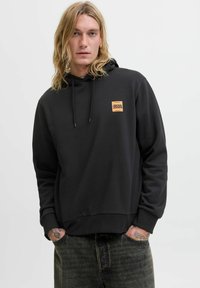 Szőke, tetoválásokkal rendelkező személy fekete kapucnis pulóverben, narancssárga Jack & Jones logóval, kezét zsebében tartva, egy fehér háttér előtt áll.