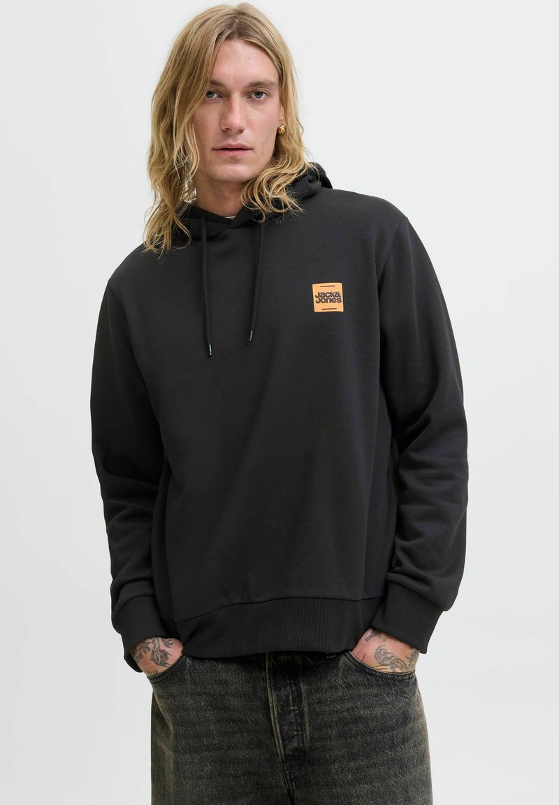 Szőke, tetoválásokkal rendelkező személy fekete kapucnis pulóverben, narancssárga Jack & Jones logóval, kezét zsebében tartva, egy fehér háttér előtt áll.