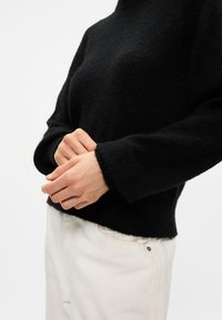Black wool sweater with a high neckline and long sleeves, featuring a soft, textured finish. Worn with light-colored pants.