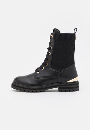 Black ankle boot with a smooth leather toe, textured knitted upper, gold eyelets, and a metallic accent on the heel. Thick black rubber sole.