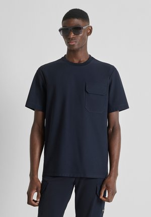 Man met zwarte zonnebril, een marineblauw shirt met korte mouwen en een borstzak, en bijpassende marineblauwe broek tegen een grijze achtergrond.