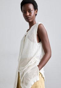 Gilet senza maniche in crema, con una texture a rete aperta, bottoni frontali e una vestibilità rilassata, indossato sopra una maglia leggera.