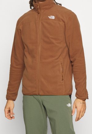 Mann trägt eine braune Fleecejacke mit Reißverschluss und grüne Hose mit The North Face Logos auf Brust und Oberschenkel.