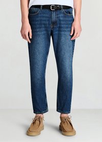 Jean en denim bleu foncé à jambes droites, texture délavée et taille mi-haute, associé à une ceinture noire et des mocassins marron clair.
