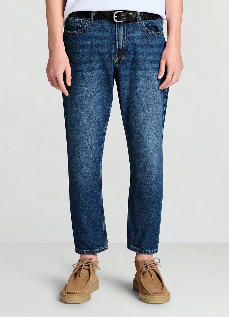 Jean en denim bleu foncé à jambes droites, texture délavée et taille mi-haute, associé à une ceinture noire et des mocassins marron clair.
