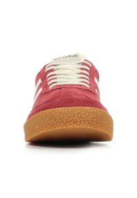 Zapatilla de ante roja con rayas laterales blancas, cordones beige y suela de goma texturizada. Punta redondeada y diseño plano. La parte superior tiene un acabado suave.