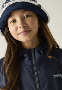 Joven chica con cabello largo y castaño que lleva un sombrero tipo bucket azul marino y una chaqueta Regatta azul marino con cremallera, sonriendo ligeramente sobre un fondo liso.