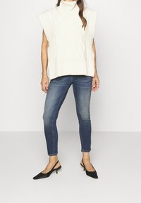 Poncho de cuello alto de punto grueso con patrones de trenza, combinado con jeans ajustados de color azul oscuro y tacones de slingback negros con punta afilada.
