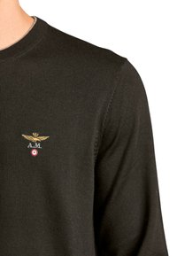 Maglione nero lavorato a maglia con scollo rotondo. Presenta un emblema alato ricamato in oro e le iniziali "A.M." sopra un punto rosso.