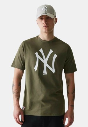 Hombre con camiseta verde oliva de los New York Yankees y gorra gris claro de los Yankees, de pie frente a un fondo liso con expresión neutral.