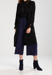 Manteau long noir avec des poignets en dentelle, chemise noire à col, et culottes larges bleu marine. Complété par des bottines à la cheville bleu marine. Texture lisse tout au long.