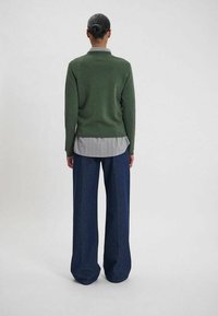 Un pull en tricot vert avec un col, superposé sur une chemise gris rayé et associé à un pantalon large bleu foncé. Fond uni.