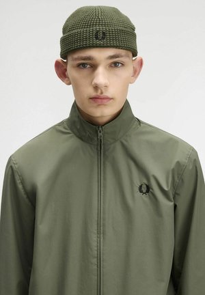 Giacca verde oliva con zip e colletto alto, con logo nero sul petto; il modello indossa un berretto in maglia testurizzata abbinato.