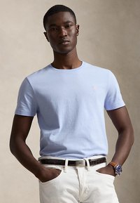 Young man wearing a light blue short-sleeve T-shirt, white pants, a black belt, and a wristwatch, standing with hands in pockets.