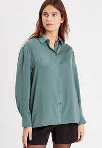 Chemise vert pâle à boutons, en tissu lisse, dotée d'un col pointu et de manches longues avec des poignets à boutons.