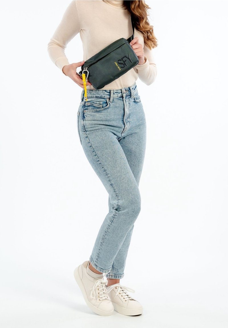 Grön tygmidjeväska med svarta detaljer, gul dragkedja och logotyp. Bärs med ljusblå jeans och vita sneakers. Enkelt design.