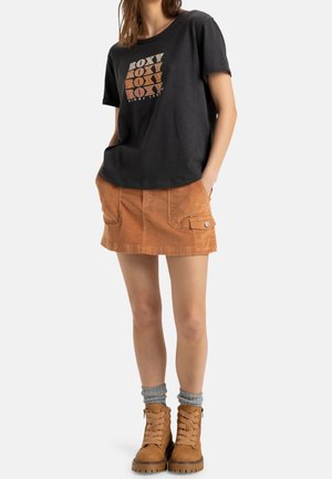 Kobieta ubrana w czarną koszulkę z nadrukiem "ROXY", brązową spódniczkę mini z sztruksu z kieszeniami, szare skarpetki i beżowe sznurowane buty, ręce w kieszeniach.