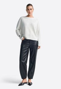 Weißer strukturierter Strickpullover mit weitem Ausschnitt, kombiniert mit schwarzen Kunstlederhosen mit lockerer Passform und umgeschlagenen Knöcheln. Schwarze verzierten Mules.