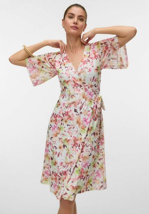 Femme ajustant le décolleté d'une robe portefeuille fleurie mi-longue avec des fleurs roses, orange et vertes sur fond blanc.