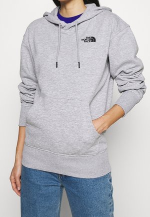 Persoon draagt een lichtgrijze The North Face hoodie met voorzak en trekkoorden, gecombineerd met blauwe jeans, hand in de zak.