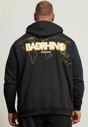 Mann, von hinten gesehen, trägt schwarzen Hoodie mit gelb umrandeter Weltkarte und dem Text „BADRHINO WORLDWIDE“ auf dem Rücken.