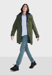 Parka impermeabile verde con foderato interno blu navy, tasca a marsupio e cappuccio regolabile, indossata sopra una polo azzurra chiara e pantaloni teal. Scarpe nere.