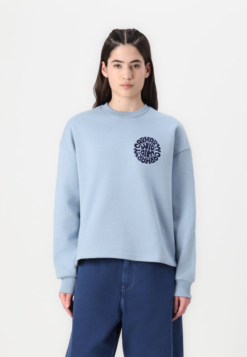 Helles blaues Sweatshirt mit gerippten Bündchen und Kragen, das ein kreisförmiges navy blaues Grafikdesign auf der Brust aufweist. Kombiniert mit navy blauen Hosen.