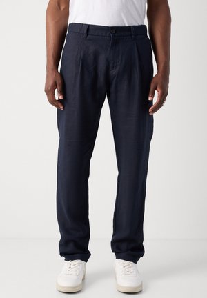 Mann trägt marineblaue plissierte Hose, weiße Turnschuhe und ein weißes T-Shirt, steht vor einem schlichten hellen Hintergrund.