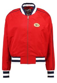 Czerwona kurtka bomberka z zamkiem z przodu, mankiety w białe i granatowe paski oraz okrągła naszywka z napisem "Miłego dnia." Gładka faktura materiału.