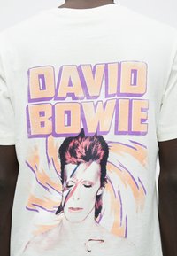 Άτομο φοράει λευκή κοντομάνικη μπλούζα με μεγάλη πολύχρωμη γραφική απεικόνιση του προσώπου του Ντέιβιντ Μπόουι, με κόκκινο και μπλε μακιγιάζ σε μορφή αστραπής, και έντονο παστέλ κείμενο "ΝΤΕΪΒΙΝΤ ΜΠΟΟΥΙ".