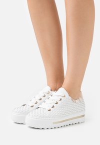 Legs wearing white woven sneakers with thick soles and gold eyelets, standing on a white background.