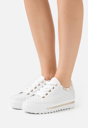 Gambe che indossano sneaker bianche intrecciate con suole spesse e occhielli dorati, in piedi su uno sfondo bianco.
