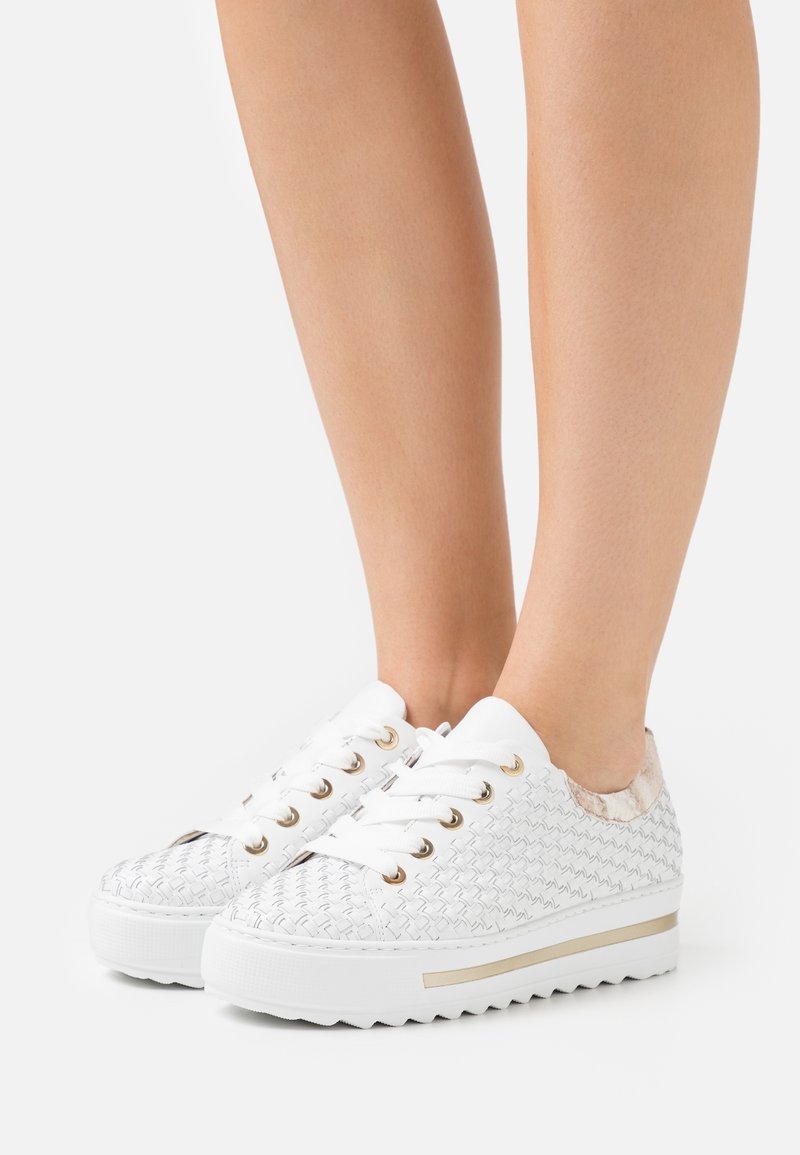 Legs wearing white woven sneakers with thick soles and gold eyelets, standing on a white background.