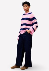 Personne debout les mains dans les poches, portant un pull rayé rose et bleu marine, un pantalon large sombre et des chaussures marron sur fond blanc.